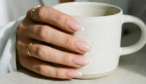 A close-up photograph of a hand with almond-shaped nails featuring a barely-there pink nude base and razor-thin white Micro French tips. The hand, wearing a delicate gold ring, holds a minimalist speckled ceramic coffee cup under natural light.