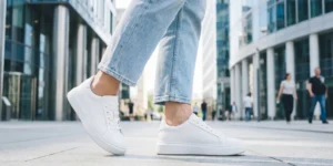 A low-angle close-up shot of a person’s feet walking on clean city pavement. They are wearing pristine, minimalist white leather sneakers with thick soles and light-wash cropped jeans. Modern glass office buildings are blurred in the background.