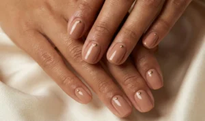Hands resting on beige silk fabric, showing almond-shaped nails with a rich, glossy nude polish that complements the skin tone. A single, tiny gold micro-stud is positioned precisely at the base of each nail.