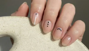 A close-up of a hand resting on speckled ceramic, showcasing natural oval nails with a nude base. Each nail has a different minimalist design: a black dot with a white line, three black dots, a small black curve, and a tiny white starburst.