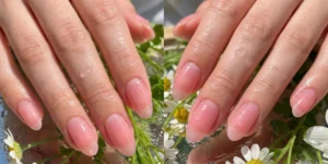 A diptych showing oval nails with a sheer, glossy "jelly" pink tint that appears wet. The hands are surrounded by fresh chamomile flowers, greenery, and water droplets.