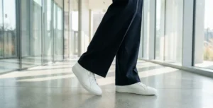A low-angle shot focusing on feet wearing clean minimalist white leather sneakers and dark wide-leg trousers walking on a polished concrete floor.