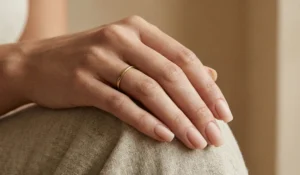 A close-up photograph of a hand with medium-length squoval nails painted in a barely-there matte nude polish that matches the skin tone. A simple thin gold band ring is on the ring finger against a blurred warm beige background.