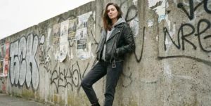 An edgy shot of a woman wearing a worn black leather moto jacket over a hoodie, leaning against a concrete wall covered in graffiti.