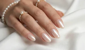 A luxurious shot of a hand wearing pearl jewelry resting on white silk. The oval nails have a sheer base topped with a highly reflective, glowing iridescent pearl glaze.