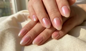 Oval nails with a highly reflective, pearlescent chrome finish that shifts between pink and white, catching the sunlight while resting on a champagne silk fabric.