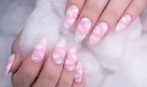 Long, almond-shaped nails resting on fluffy white material. The nail art features sponged soft white and cotton-candy pink shades creating a cloud texture, with tiny, subtle white hearts hidden within the pattern.