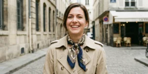 A portrait of a smiling woman wearing a beige trench coat and a patterned silk scarf tied around her neck, standing on an old stone street.
