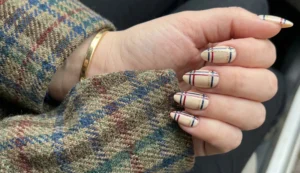 A close-up of almond-shaped nails with a beige base, featuring thin, precise plaid lines in navy blue, deep red, and gold glitter. The hand rests on a tweed jacket.