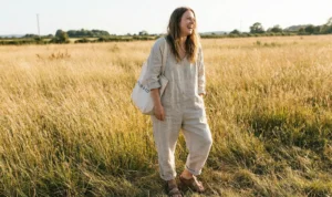 A smiling woman wearing a loose beige linen jumpsuit and holding a 'FIELD & CO.' tote bag, standing in a sunlit field of tall golden grass.