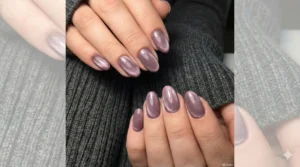 Hands wearing a dark grey ribbed knit sweater, featuring oval-shaped nails painted in a dusty mauve magnetic polish. The light reflection on the nails creates a shifting, shimmering "velvet" texture effect.