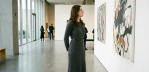 A woman wearing a long, charcoal grey rib-knit dress stands in a modern art gallery, looking at a large abstract painting on the wall.