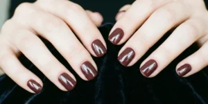 Short, squoval nails painted in a deep, dark oxblood or brownish-red color with an extremely high-gloss, glass-like finish, contrasting against fair skin and dark velvet.