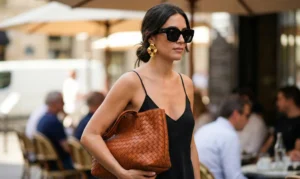 A stylish woman wearing large sunglasses and gold earrings, clutching an oversized woven brown leather tote bag under her arm on an outdoor cafe patio.