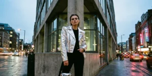 A woman wearing a metallic silver motorcycle jacket and holding a small, geometric reflective handbag, standing on a wet city street corner at dusk.
