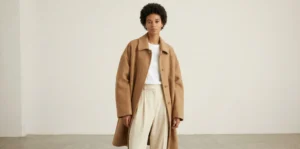 A woman with short natural hair standing in a minimalist studio, wearing an oversized camel wool coat, a white t-shirt, and wide-leg beige trousers.