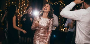 A smiling woman in a sparkling dress holds a glass of champagne on a dance floor.