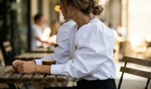 Side view of a woman sitting at an outdoor cafe table, wearing a fashionable white blouse with large, dramatic puff sleeves