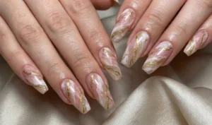 Close-up of long, coffin-shaped nails with a luxury marble effect. Swirls of soft pink, white, and translucent beige create a stone look, accented by fine veins of metallic gold leaf threaded through the pattern, mimicking Kintsugi cracks.