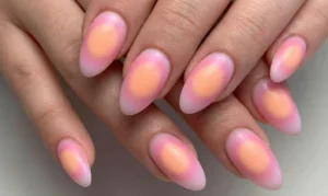 A close-up photograph of almond-shaped nails with a glowing "aura" gradient design. A central circle of warm peach color fades seamlessly into a cool pink glow toward the edges of the nails.