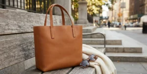 Lifestyle product shot of a tan leather tote bag resting on a wooden bench with sunglasses and a cream scarf, in an outdoor urban setting.