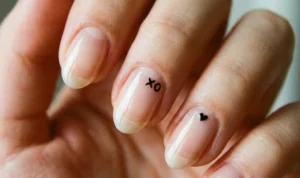 A detailed macro shot of natural nails with a clear glossy base. A tiny black minimalist "xo" is painted near the cuticle of one nail, and a microscopic black heart dot is on another.