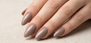 A clean, professional photograph focusing on a graceful hand resting on beige linen. The nails are perfectly sculpted in a long almond shape and painted with a solid, classic glossy taupe polish.