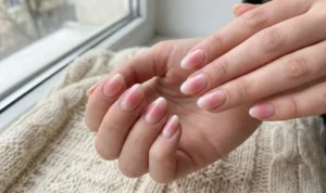Oval nails with a "blush" gradient effect, featuring a concentrated pink color in the center fading into a pale nude tip with a glossy finish, resting on a cream knitted surface.