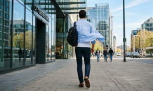A dynamic street shot of a man walking quickly away from the camera, wearing dark wash slim jeans, a white tee, and a light blue Oxford shirt completely open and flowing in the wind. Brown loafers are visible.