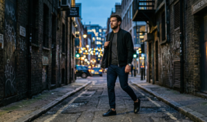 A candid, moody street portrait of a man walking through a gritty urban alleyway at twilight, wearing dark navy slim chinos, a charcoal grey tee, and a sleek black bomber jacket.