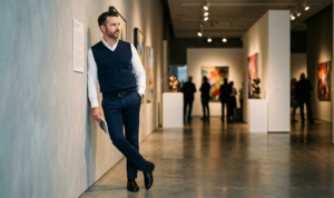 A sophisticated portrait of a man in navy slim-fit trousers and a white Oxford shirt, layered with a polished navy blue knit vest, looking intelligent inside a gallery with focused spotlights and blurred art.