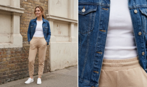 A full-length candid photograph of a smiling young woman with dark blonde wavy hair walking confidently towards the camera along a bustling urban sidewalk in London. She is mid-stride, wearing an oversized, distressed charcoal grey graphic t-shirt with a white and red band logo loosely front-tucked into black high-waisted biker shorts. She has a small black leather crossbody bag slung over her left shoulder and wears white ribbed athletic socks with chunky white "dad" sneakers. The background features historical brick buildings, a coffee shop with patrons, and other pedestrians under diffused daylight.