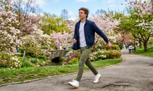 An action portrait of a man walking energetically through a London park with blooming trees, wearing slim olive green chinos, a plain white tee, and a crisp navy Harrington jacket.
