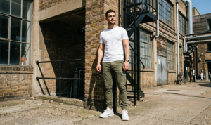 A rugged street portrait of a man standing near industrial-chic architecture, wearing tailored olive green cargo trousers, a fitted white tee, and chunky white 'dad' trainers.