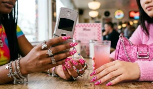 Two pairs of hands displaying vibrant hot pink manicures heavily adorned with sparkling Y2K-inspired rhinestones and crystals while holding a retro flip phone.