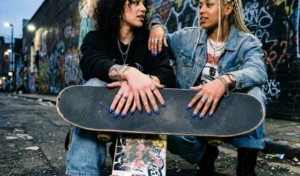 Close-up of street-style almond nails painted with electric cobalt blue flame designs on a clear base, resting on a skateboard.