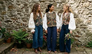 Three women leaning against a textured stone wall wearing richly embroidered tapestry vests layered over billowy white poet blouses and wide-leg trousers for a bohemian Renaissance flair.